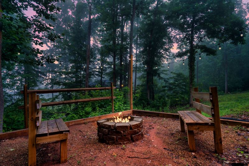Stone fire pit burning at dusk between two wooden benches under string lights